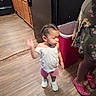 toddler, child, indoor, kitchen, wooden_floor, cabinet, trash_bin, person, clothing, footwear, braided_hair, pink_shorts, white_shirt, crocs, floral_dress, motion_blur, floor, appliance, home, adult