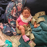 Kinsliegh is registered to the contest to win money with this photo: baby, dollar_bills, pink_clothing, bib, blanket, bed, smile, happy, infant, money, currency, indoor, cute, child, sitting, playful, soft_texture, fabric, cozy, wrinkled_clothing