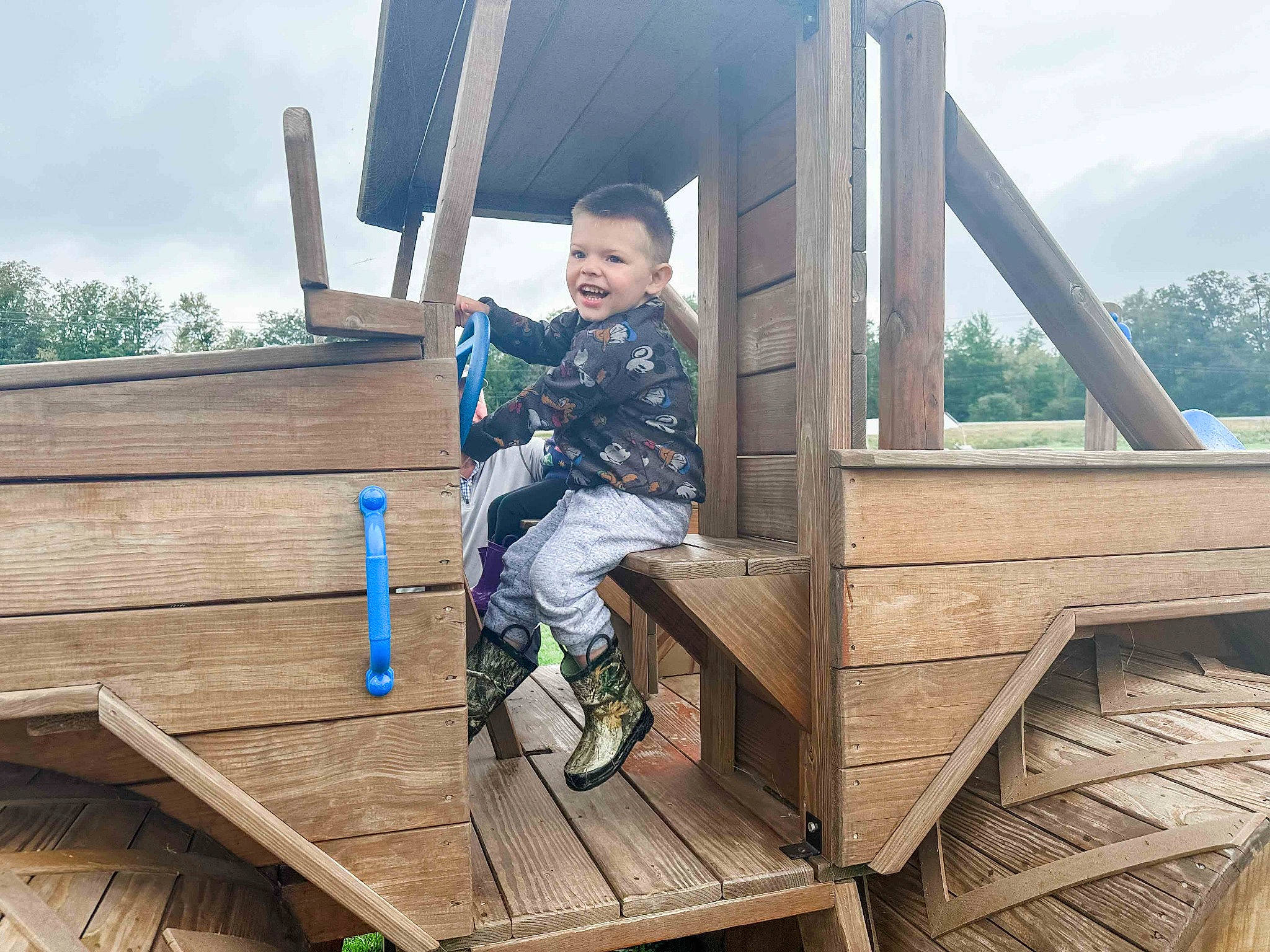 Graeson is registered to the contest to win money with this photo: cloud, fun, grass, hardwood, house, jacket, joy, leg, leisure, outdoor_shoe, person, plant, recreation, shoe, shorts, sitting, sky, smile, sneakers, toddler