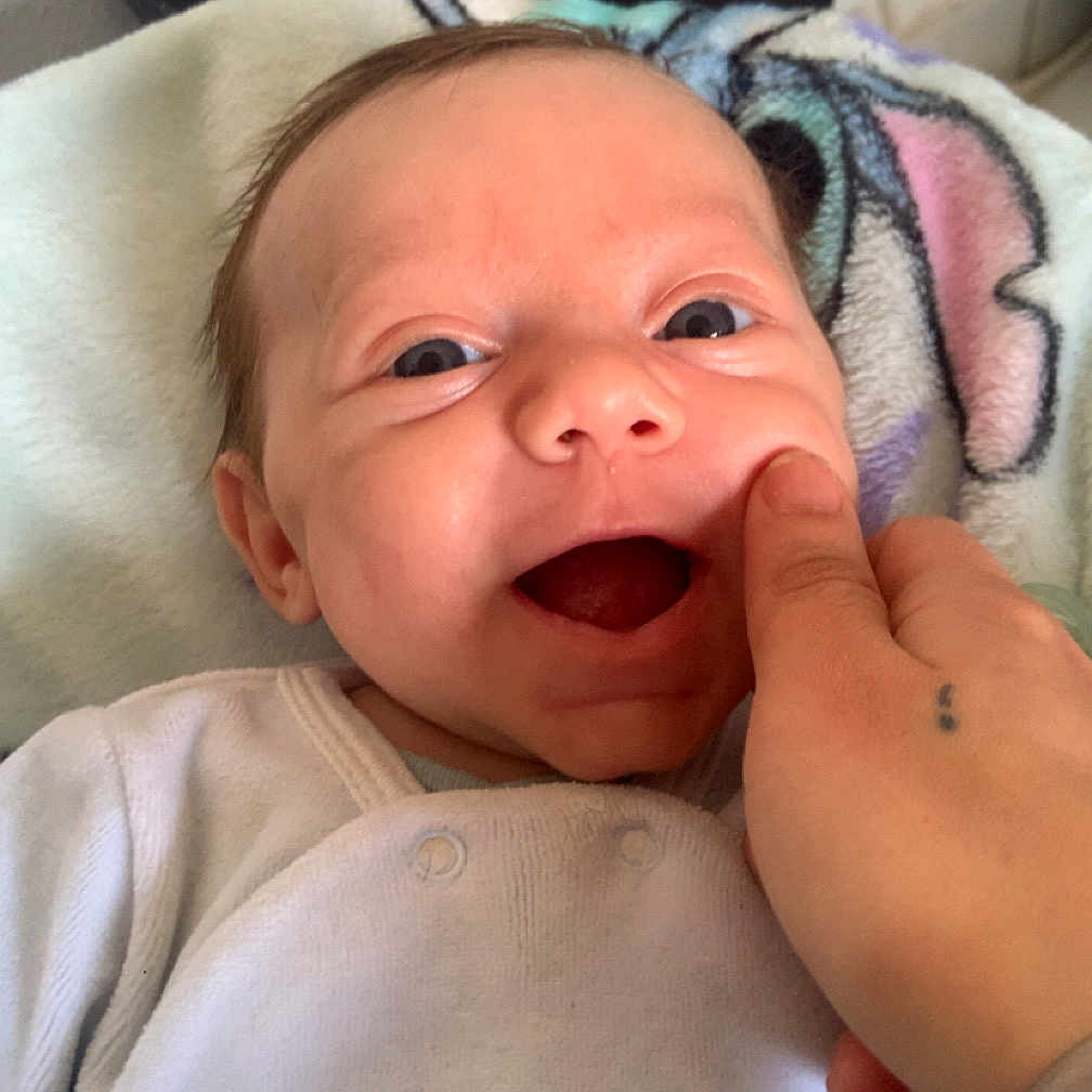 Maël participe au concours pour gagner de l'argent avec cette photo : baby, blanket, cheek, closeup, clothing, comfort, cute, expression, face, fingertip, hand, happy, indoor, infant, onesie, person, skin, smiling, tattoo, young_child