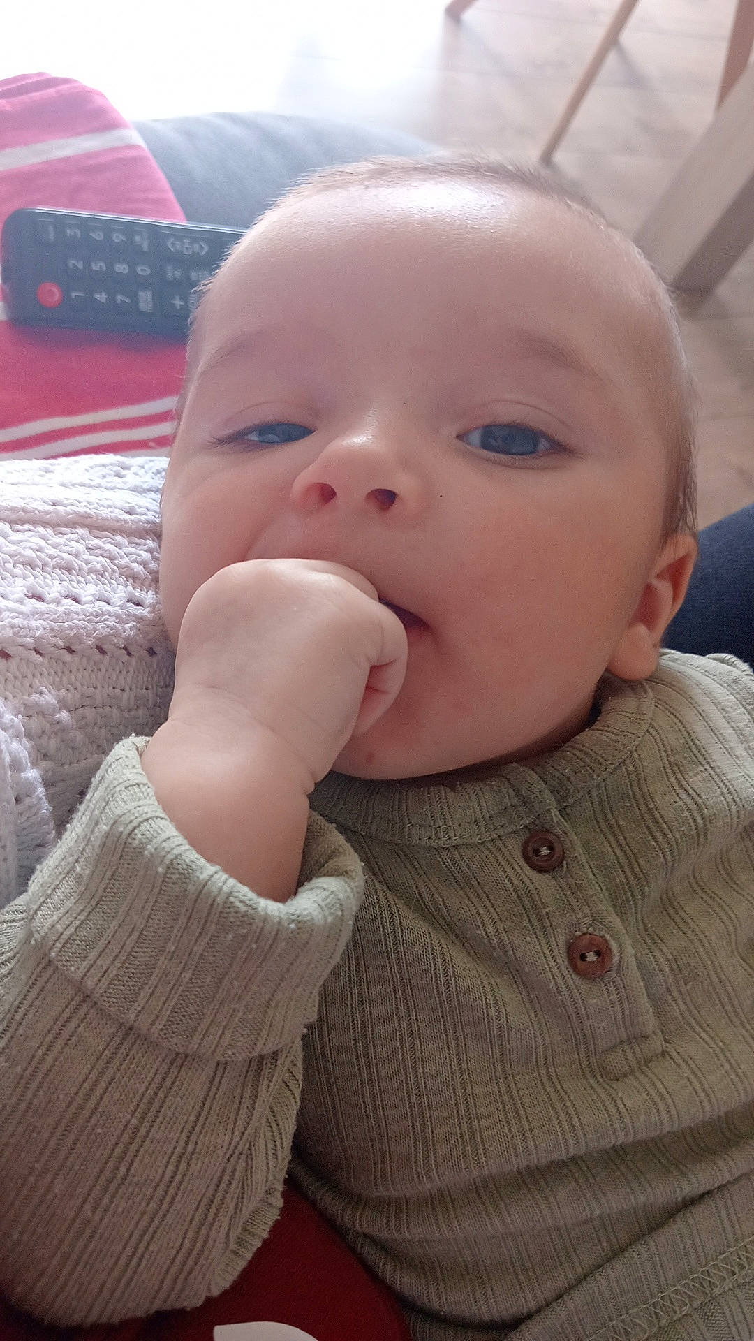 baby, blanket, blue_eyes, buttoned_clothing, child, close_up, couch, cozy, face, furniture, hand, indoor, infant, knitted, person, portrait, relaxed, remote_control, skin, sweater