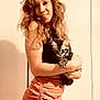 woman, cat, kitten, smiling, curly_hair, holding, indoor, portrait, casual_clothing, skirt, black_top, light_background, feline, pet, happy, cute, young_adult, warm_lighting, friendly, cozy