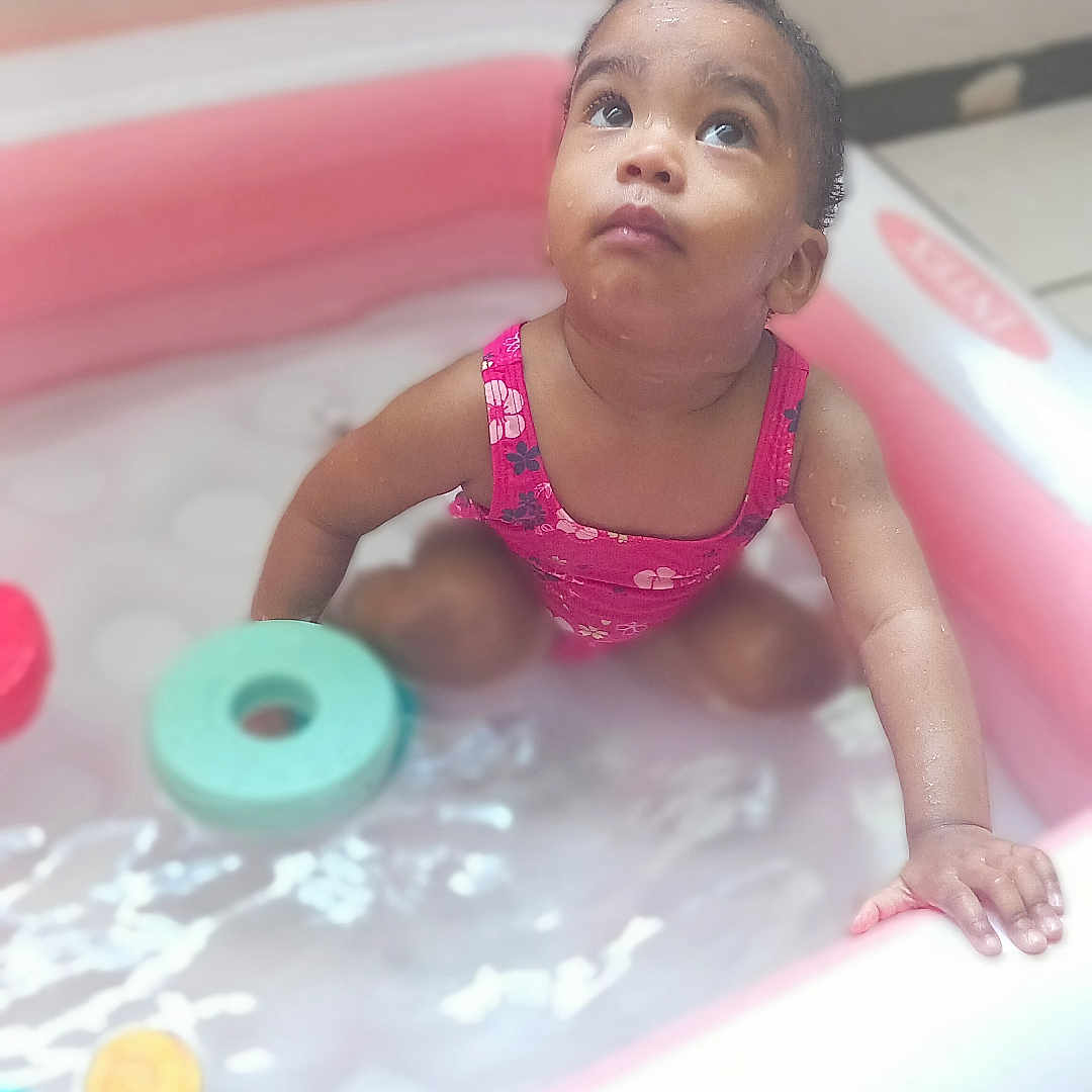 Leïna Kiara participe au concours pour gagner de l'argent avec cette photo : bath_time, bright, child, curious, cute, face, floral_pattern, hand, indoor, inflatable_pool, looking_up, pink, playing, sitting, skin, small_pool, swimsuit, toddler, toy, water