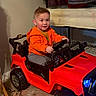 Shay joined the competition — help win amazing prizes! toddler, child, toy_car, indoor, orange_hoodie, red_car, car_toy, happy, smiling, steering_wheel, clothing, floor, jacket, shelf, furniture, person, play, cute, car_wheels, room