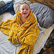 Kai joined the competition — help win amazing prizes! baby, child, blanket, bed, pillow, smile, happy, indoor, yellow, patterned_blanket, person, cute, cozy, wrapped, head, face, young_child, warm, comfort, home
