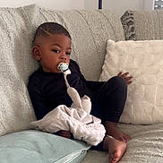 Idriss participe au concours pour gagner de l'argent avec cette photo : child, toddler, pacifier, blanket, couch, pillow, indoor, relaxed, black_clothing, barefoot, soft_texture, home, infant, seated, comfort, young_child, curly_hair, cozy, furniture, portrait