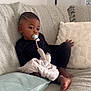 child, toddler, pacifier, blanket, couch, pillow, indoor, relaxed, black_clothing, barefoot, soft_texture, home, infant, seated, comfort, young_child, curly_hair, cozy, furniture, portrait