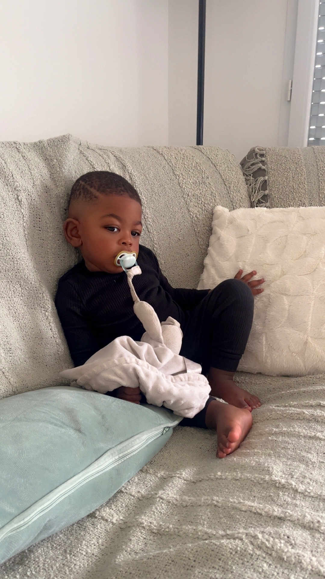 Idriss participe au concours pour gagner de l'argent avec cette photo : child, toddler, pacifier, blanket, couch, pillow, indoor, relaxed, black_clothing, barefoot, soft_texture, home, infant, seated, comfort, young_child, curly_hair, cozy, furniture, portrait