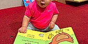 Khalesei is registered to the contest to win money with this photo: toddler, child, book, reading, pink_shirt, bow, carpet, red_carpet, bookshelf, children_books, board_book, animal_illustration, indoor, toy_store, curious, sitting, face, person, early_learning, playful