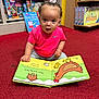 toddler, child, book, reading, pink_shirt, bow, carpet, red_carpet, bookshelf, children_books, board_book, animal_illustration, indoor, toy_store, curious, sitting, face, person, early_learning, playful