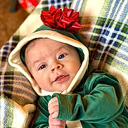 Nicholas is registered to the contest to win money with this photo: baby, infant, face, hood, bow, green_clothing, plaid_blanket, smile, hand, blue_eyes, cute, indoors, portrait, child, soft_texture, button, warm_clothing, closeup, relaxed, lying_down