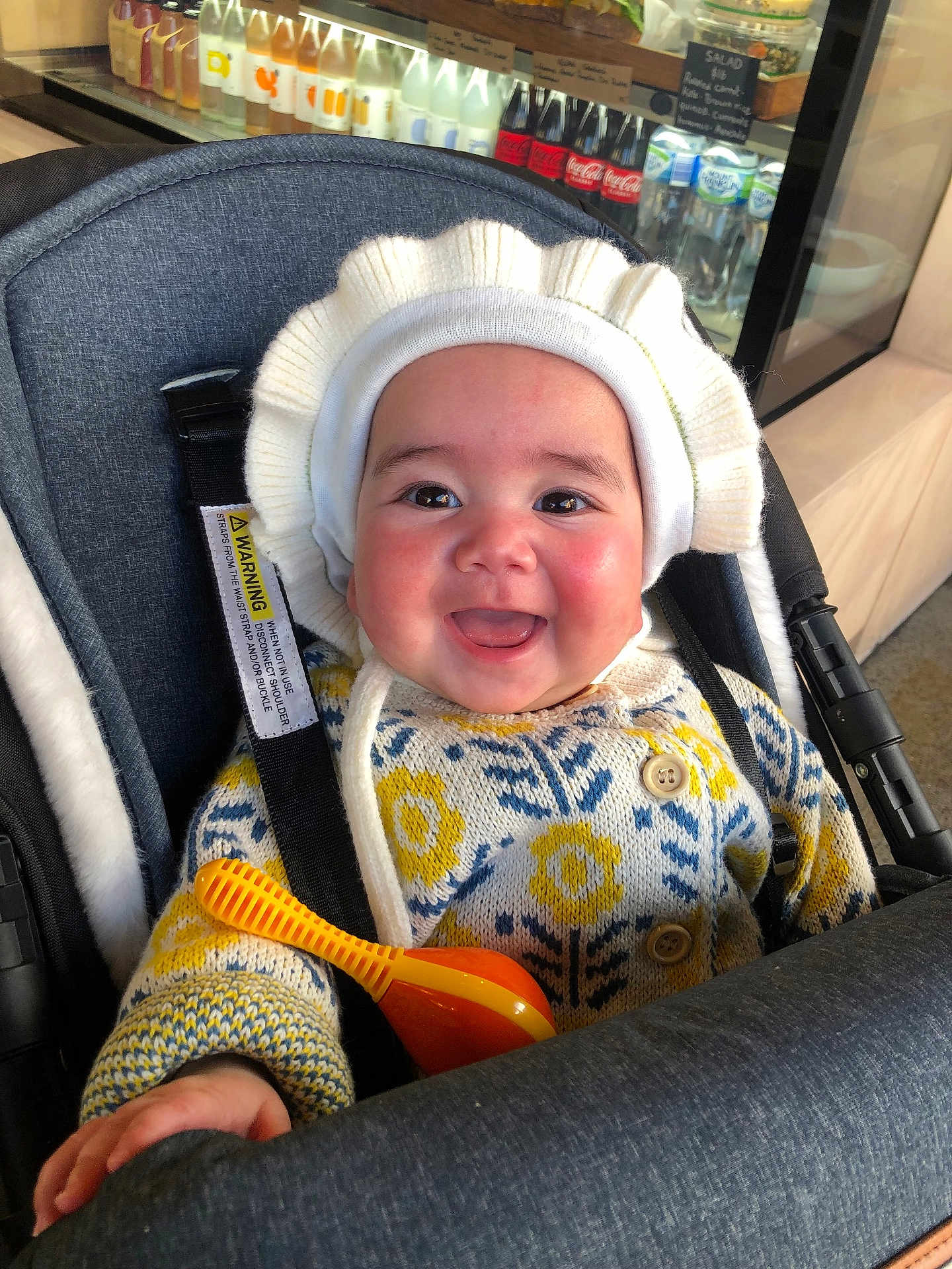 Daniela is registered to the contest to win money with this photo: baby, smiling, stroller, hat, sweater, toy, indoor, drink_bottles, refrigerator, happy, child, face, clothing, button, seatbelt, cafe, person, cute, warm_clothing, infant