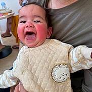 Daniela is registered to the contest to win money with this photo: baby, child, adult, laughing, smiling, happy, rosy_cheeks, quilted_clothing, lion_patch, indoor, family, holding, person, face, head, hand, background, blurred, casual_clothing, people
