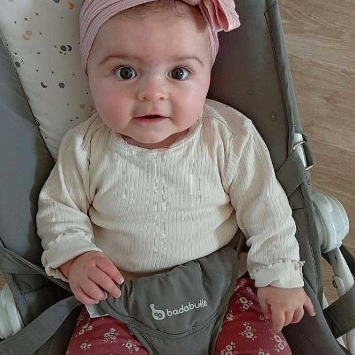 Athéna participe au concours pour gagner de l'argent avec cette photo : baby, baby_bouncer, child, clothing, comfort, cream_top, cute, expression, face, flooring, floral_pants, headband, indoor, infant, infant_clothing, looking_at_camera, pink_bow, portrait, seated, wide_eyes