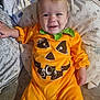 baby, child, pumpkin_costume, orange_clothing, smiling, face, cushion, blanket, indoor, cozy, cute, holiday, festive, halloween, infant, person, apparel, head, happy, toddler