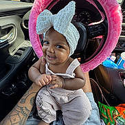 Aulelei is registered to the contest to win money with this photo: baby, child, headband, bow, smile, car_interior, steering_wheel, pink, fuzzy, tattoo, clothing, overalls, snack, seat, window, daylight, person, hand, foot, happy