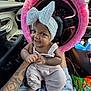 baby, child, headband, bow, smile, car_interior, steering_wheel, pink, fuzzy, tattoo, clothing, overalls, snack, seat, window, daylight, person, hand, foot, happy