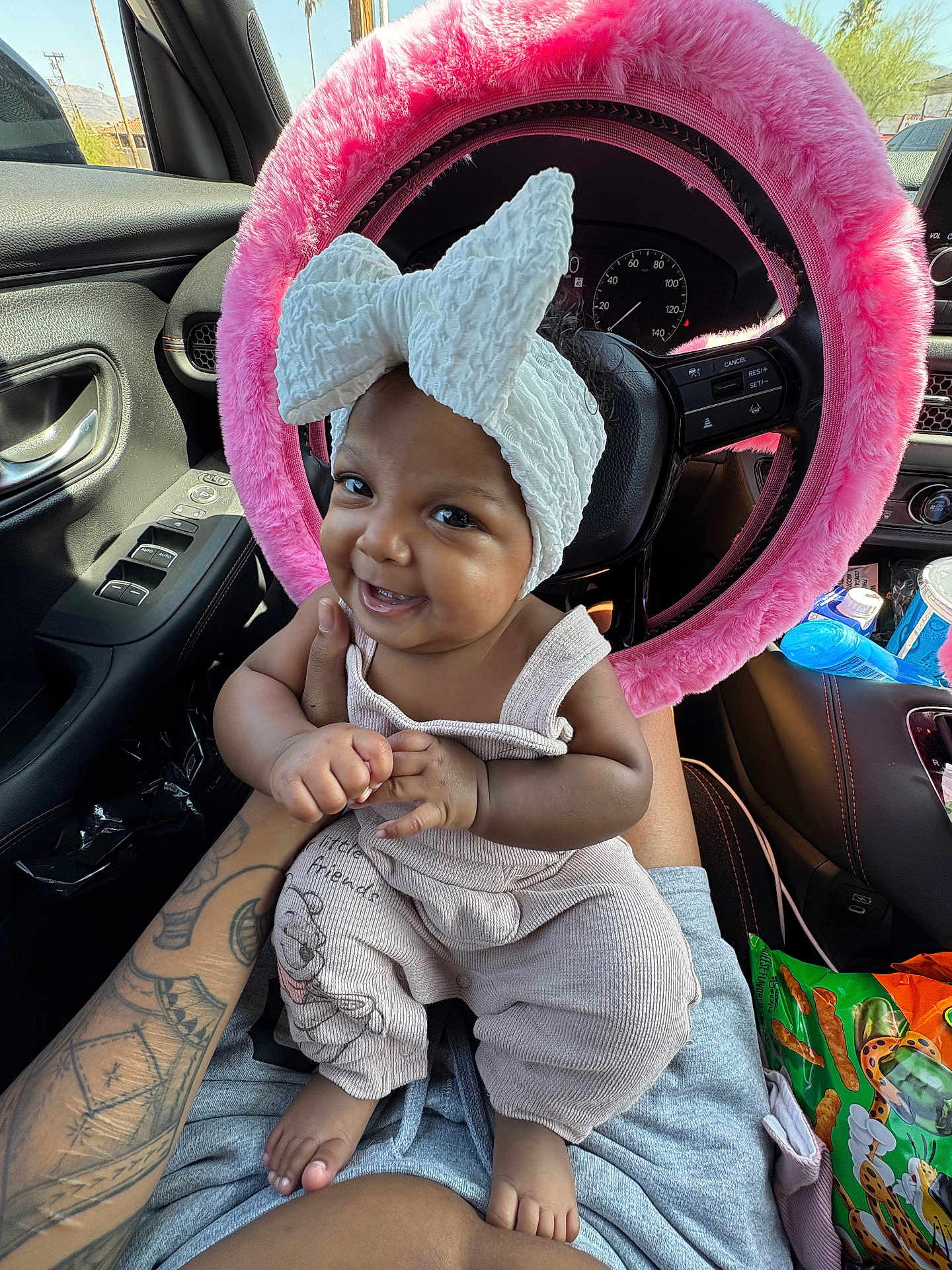 Aulelei is registered to the contest to win money with this photo: baby, child, headband, bow, smile, car_interior, steering_wheel, pink, fuzzy, tattoo, clothing, overalls, snack, seat, window, daylight, person, hand, foot, happy