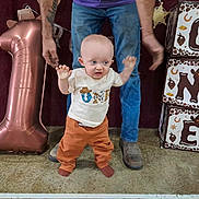 Jasper is registered to the contest to win money with this photo: baby, child, adult, jeans, balloon, number_one, decorations, floor, shoes, socks, shirt, pants, hands, celebration, party, indoor, blocks, standing, helping, person