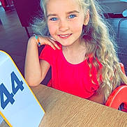 Jade a rejoint le concours — aidez-le/la à gagner de superbes lots ! child, girl, smiling, blue_eyes, long_hair, curly_hair, red_shirt, bracelet, table, restaurant, indoor, casual, number_sign, seated, person, happy, portrait, light, furniture, background