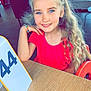 child, girl, smiling, blue_eyes, long_hair, curly_hair, red_shirt, bracelet, table, restaurant, indoor, casual, number_sign, seated, person, happy, portrait, light, furniture, background