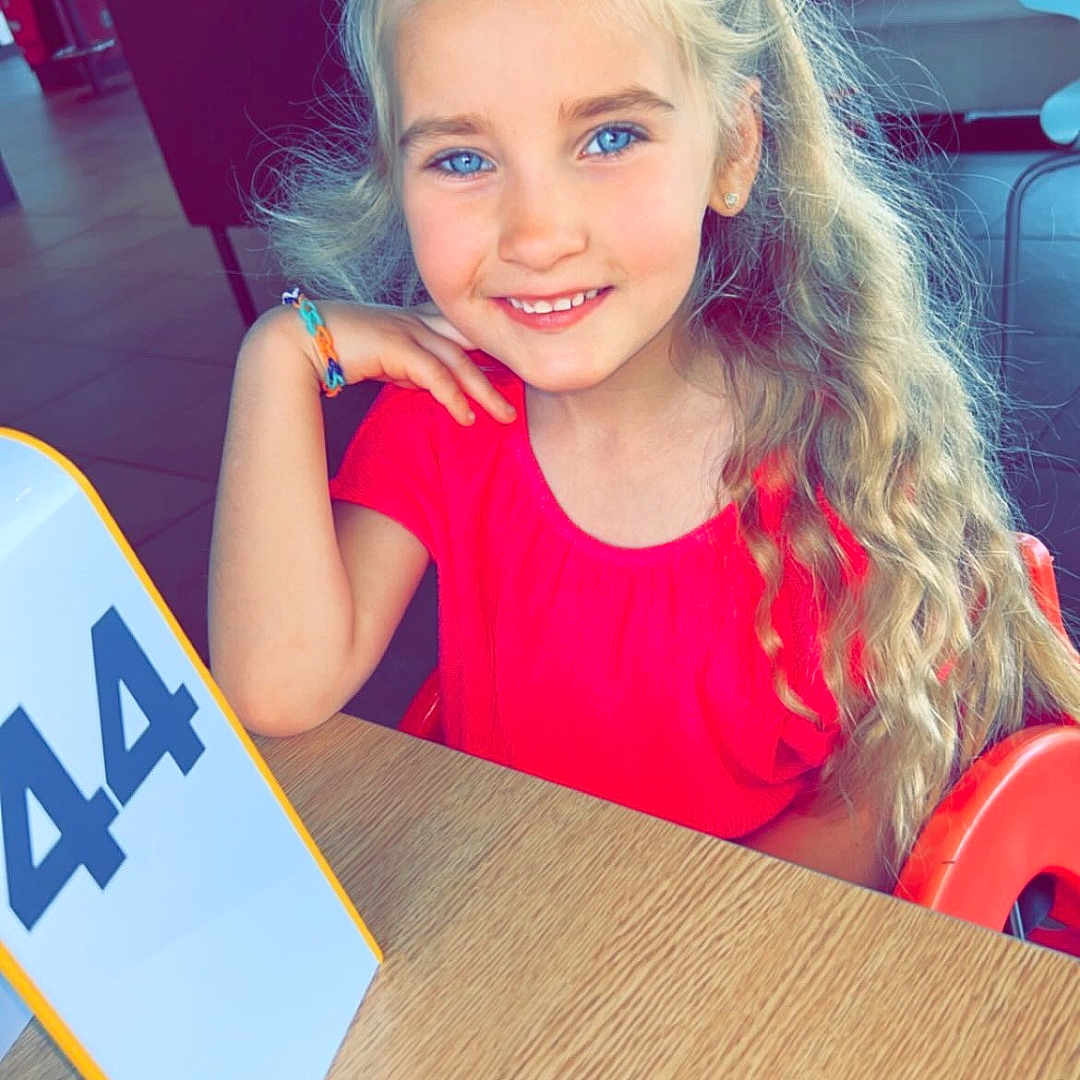 Jade a rejoint le concours — aidez-le/la à gagner de superbes lots ! background, blue_eyes, bracelet, casual, child, curly_hair, furniture, girl, happy, indoor, light, long_hair, number_sign, person, portrait, red_shirt, restaurant, seated, smiling, table