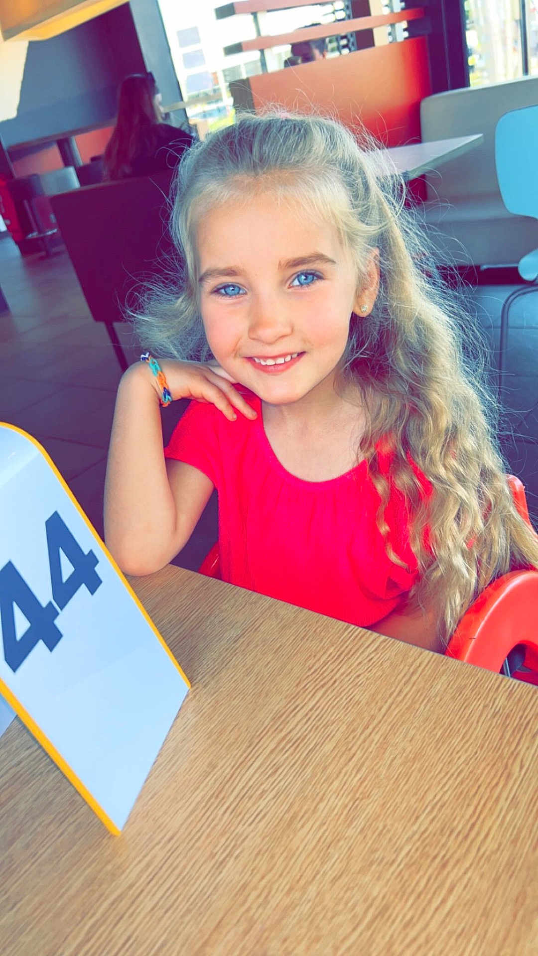 Jade a rejoint le concours — aidez-le/la à gagner de superbes lots ! child, girl, smiling, blue_eyes, long_hair, curly_hair, red_shirt, bracelet, table, restaurant, indoor, casual, number_sign, seated, person, happy, portrait, light, furniture, background