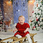 Lottie is registered to the contest to win money with this photo: baby, child, sled, red_dress, snow, christmas_tree, reindeer, holiday, winter, festive, smiling, indoor, decorations, white_tights, wood, cute, portrait, seasonal, happy, celebration