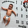 baby, blanket, bodypart, clothing, diaper, face, finger, hand, head, newborn, number, person, photography, portrait, quilt, sitting, symbol, t_shirt, text, wrist