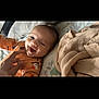 baby, smiling, infant, onesie, orange_clothing, halloween_theme, ghost_print, pumpkin_print, bed, blanket, hand, tickling, happy, lying_down, face, person, indoors, cozy, cute, playful