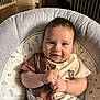 baby, infant, smiling, baby_seat, dinosaur_print, brown_clothing, beige_clothing, cushion, indoor, child, cute, happy, plush, straps, portrait, small_hands, face, eyes, hair, seat