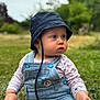 baby, child, denim_overall, bucket_hat, outdoor, grass, wooden_stick, curious, sitting, nature, greenery, portrait, cute, young_child, casual_clothing, daylight, blue_buttons, closeup, infant, expression