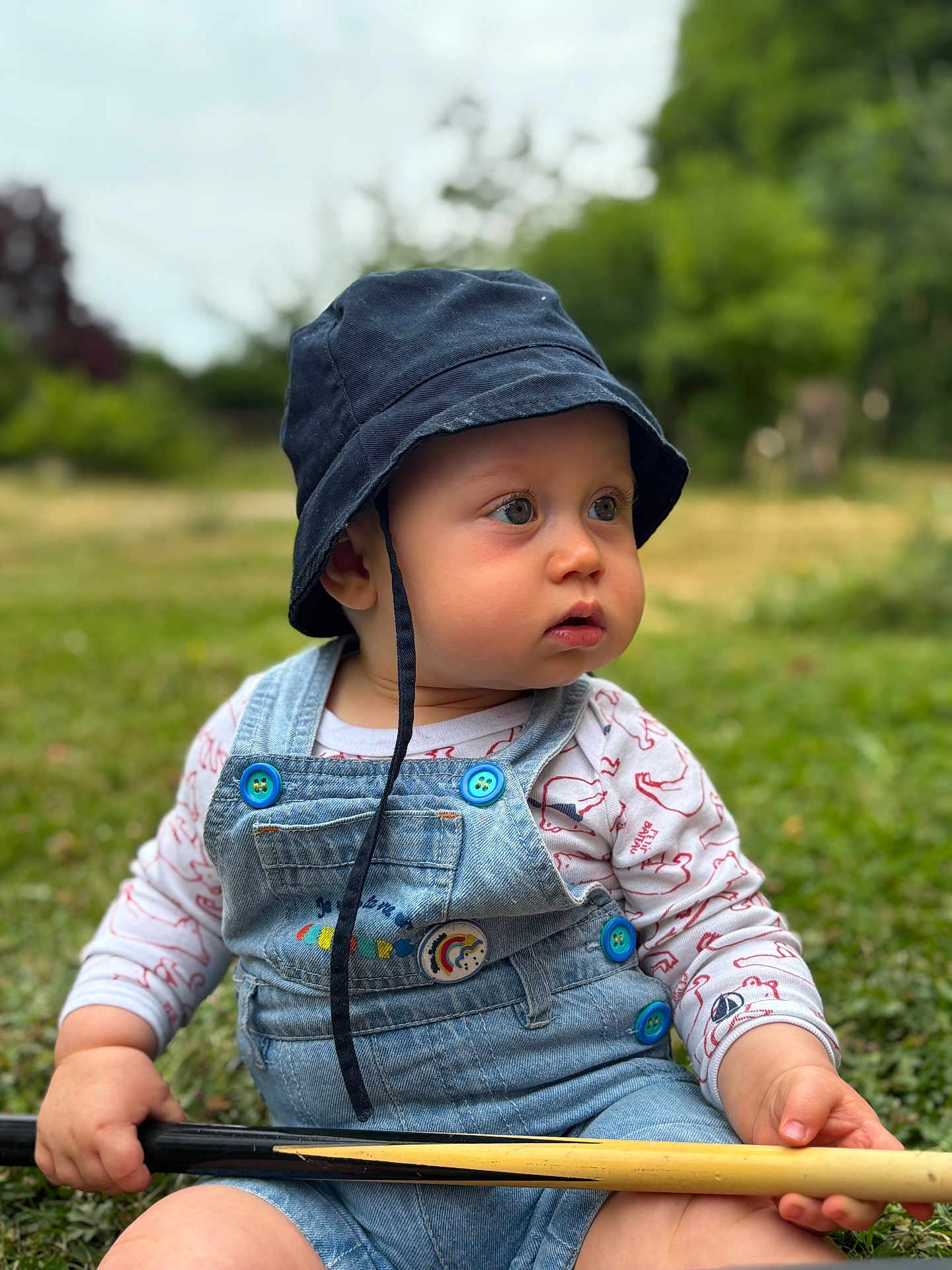 Morgan participe au concours pour gagner de l'argent avec cette photo : baby, child, denim_overall, bucket_hat, outdoor, grass, wooden_stick, curious, sitting, nature, greenery, portrait, cute, young_child, casual_clothing, daylight, blue_buttons, closeup, infant, expression