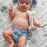 Liam participe au concours pour gagner de l'argent avec cette photo : baby, infant, crochet_hat, crochet_diaper, toy, bone_toy, lying_down, bedspread, floral_pattern, cute, child, smiling, blue, handmade, soft_toy, indoor, person, headwear, clothing, skin