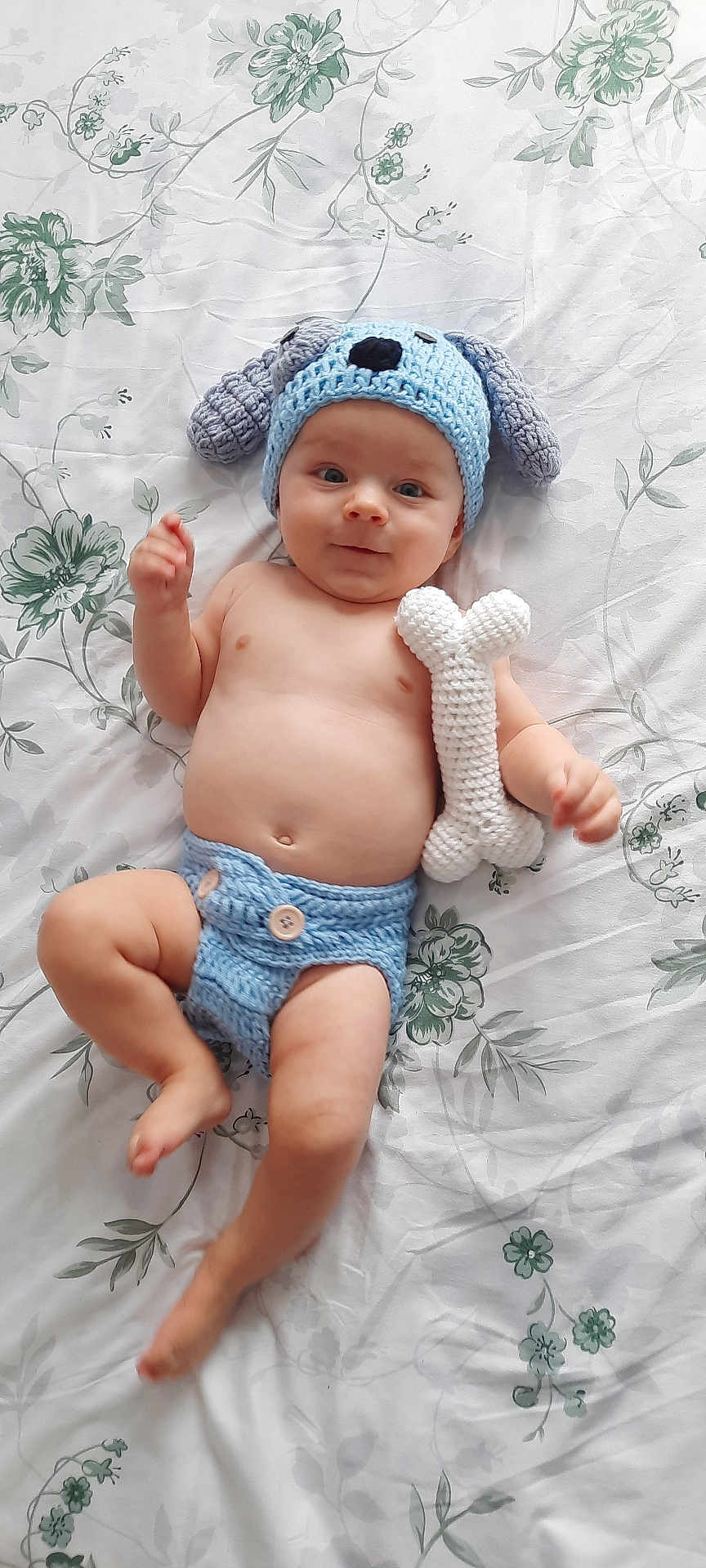 Liam participe au concours pour gagner de l'argent avec cette photo : baby, infant, crochet_hat, crochet_diaper, toy, bone_toy, lying_down, bedspread, floral_pattern, cute, child, smiling, blue, handmade, soft_toy, indoor, person, headwear, clothing, skin
