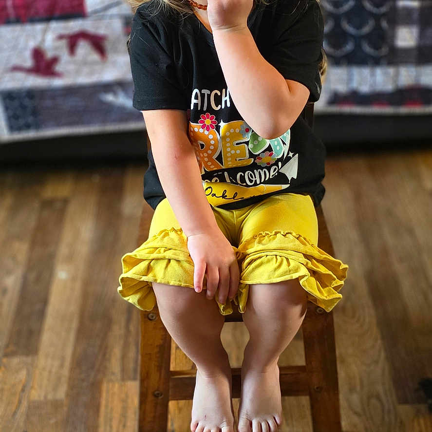 Oakley Young is registered to the contest to win money with this photo: barefoot, black_tshirt, casual, child, cute, girl, glasses, happy, home, indoor, pink_bow, playful, portrait, quilt, sitting, smiling, wood_floor, wooden_chair, yellow_skirt, young