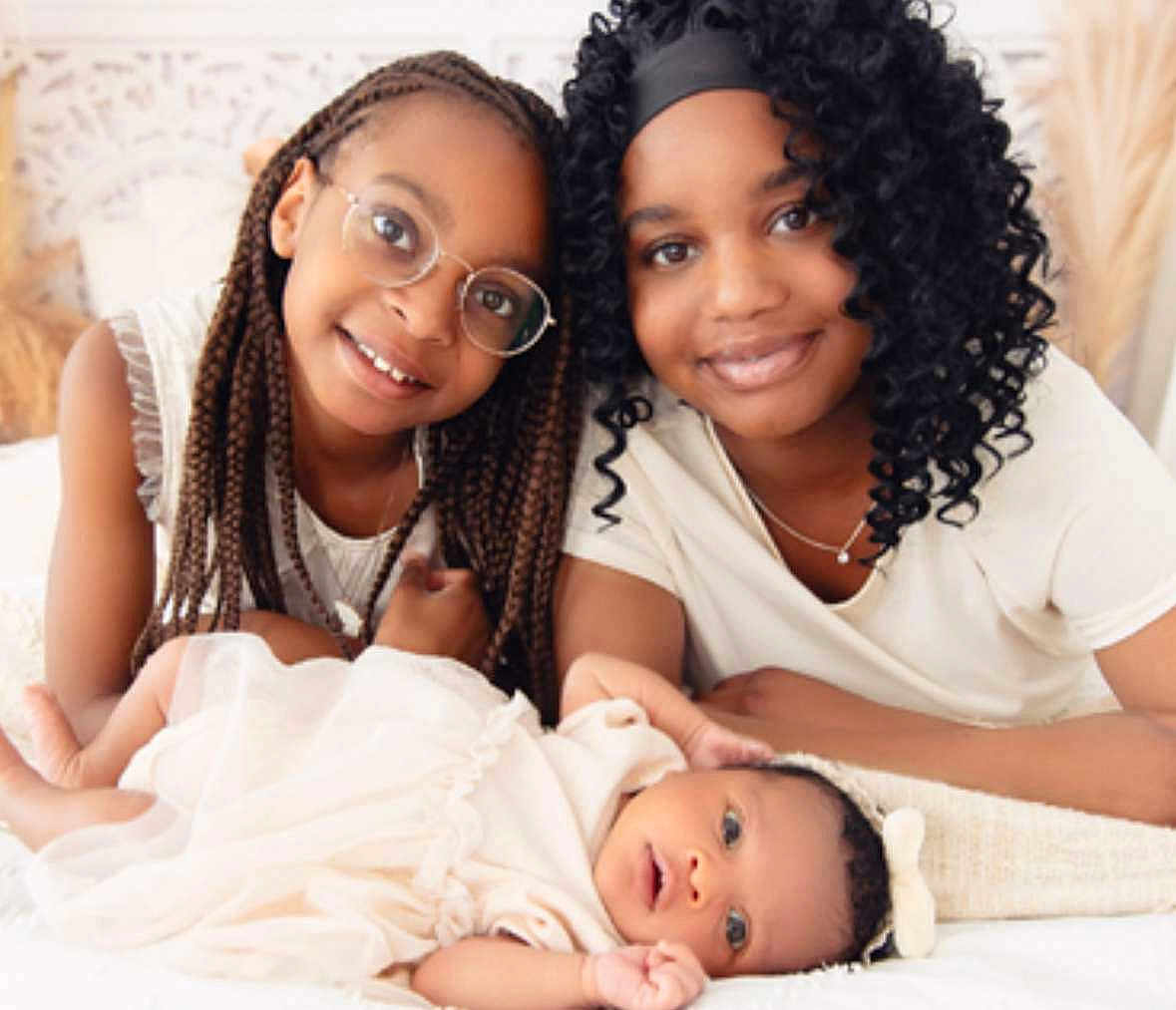 Kélyssia a rejoint le concours — aidez-le/la à gagner de superbes lots ! child, baby, smiling, glasses, braids, curly_hair, bed, white_clothing, headband, family, portrait, indoors, happy, together, people, skin_tone, candid, soft_light, blanket, cute