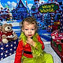 child, toddler, grinch_costume, holiday_backdrop, whoville_sign, stuffed_toy, teddy_bear, presents, festive, christmas, costume, curly_hair, blue_eyes, sitting, portrait, colorful, indoor, playful, fur_trim, gift_box