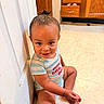 baby, casual, child, cute, floor, floor_tile, hair_bun, happy, indoor, kitchen, light_skin, onesie, person, playful, sitting, smiling, striped_clothing, toddler, wooden_cabinets, young_child