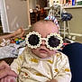 baby, sunglasses, flower_frame, indoor, person, hand, clothing, seat, home, living_room, adult_hand, cute, sun_pattern, infant, face, eyewear, holding, casual, relaxed, comfort
