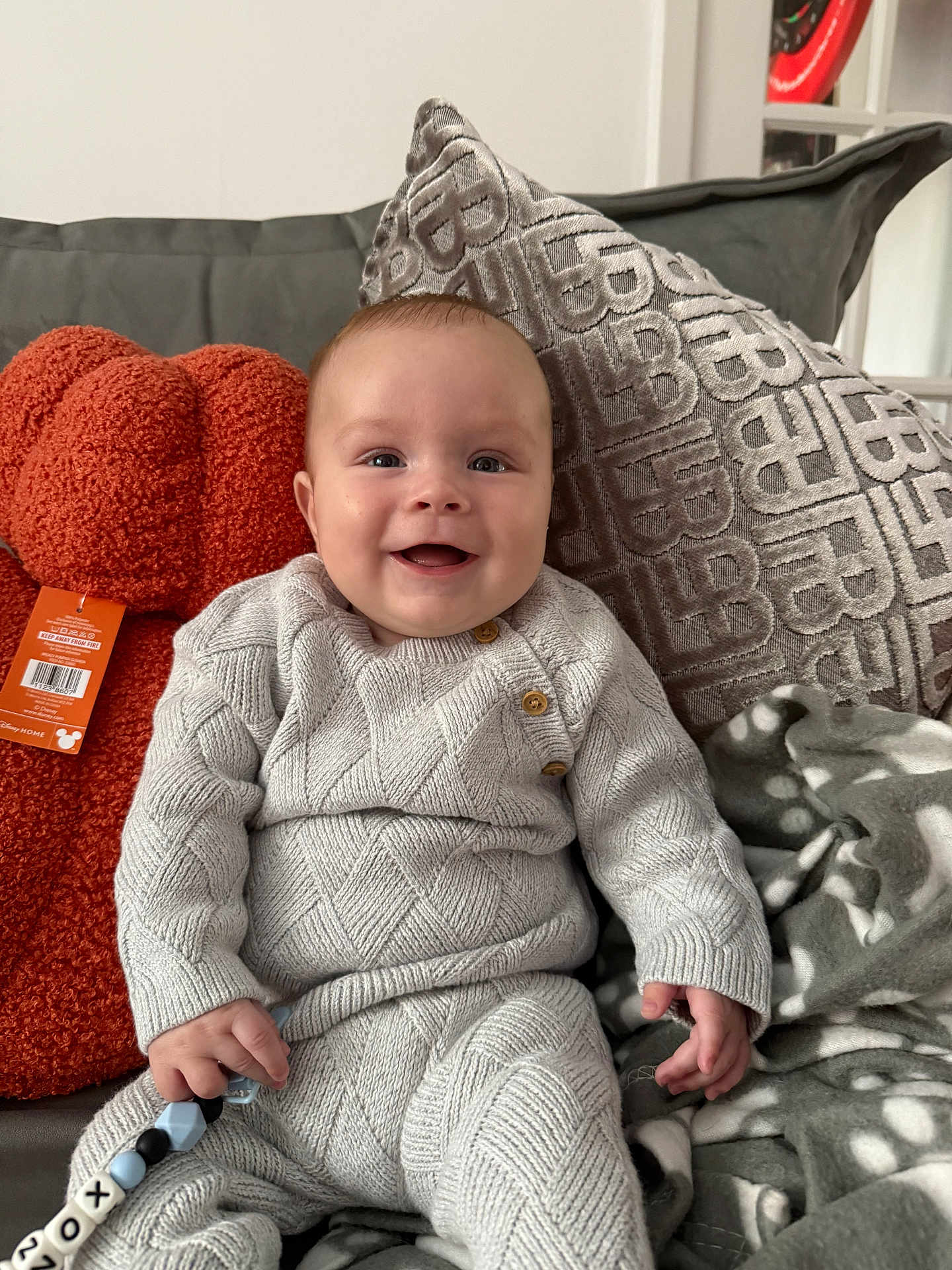 Lennox is registered to the contest to win money with this photo: baby, child, smile, knitted_clothing, couch, cushion, blanket, toy, indoor, happy, face, hand, button, pattern, plush, comfort, sitting, person, home, cozy