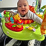 activity_jumper, baby, carpet, child, clothing, colorful, cute, floor, furniture, happy, head, indoor, infant, living_room, person, plastic, playful, seat, smiling, toys