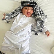 Kaïs participe au concours pour gagner de l'argent avec cette photo : baby, child, smiling, white_clothing, headscarf, lying_down, bed, happy, cute, infant, person, indoors, patterned_fabric, traditional_clothing, face, small_child, portrait, resting, cheerful, young