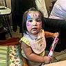 toddler, face_paint, butterfly_paint, bib, yellow_clothing, chair, cushion, table, person, indoor, curious_expression, child, pink_item, hair_tie, floor_tiles, furniture, table_leg, wall, adult, portrait