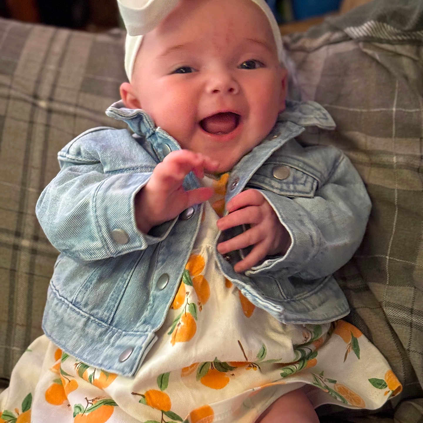 Clara joined the competition — help win amazing prizes! baby, bow, child, clothing, cozy, cute, denim_jacket, face, hands, happy, headband, indoors, infant, orange_print_dress, person, plaid_couch, portrait, sitting, smiling, young
