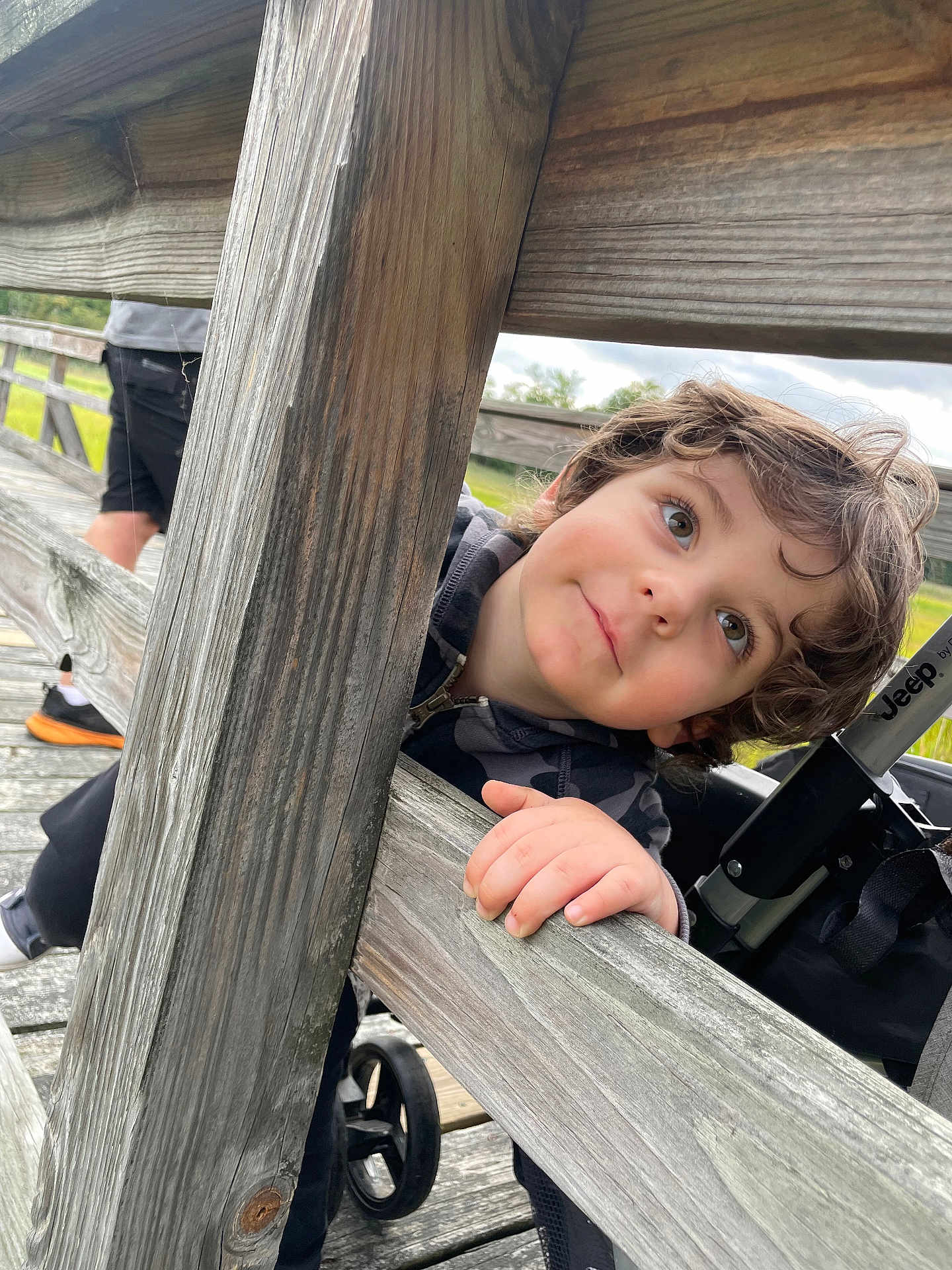 Chase is registered to the contest to win money with this photo: child, toddler, wood, bridge, hand, face, curly_hair, outdoor, stroller, person, smiling, eyes, casual, playful, railing, wooden_planks, nature, daylight, expression, clothing