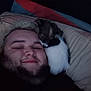 Reggie is registered to the contest to win money with this photo: person, dog, pillow, bed, smile, face, beard, hair, black_clothing, indoor, resting, snuggling, cute, pet, friendship, cozy, closeup, dark_background, comfort, sleepy