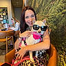 woman, dog, sunglasses, pink_harness, leopard_print_dress, smiling, cafe, table, chair, forest_mural, indoor, person, laptop, wooden_table, happy, pet, seated, casual, brown_hair, accessories