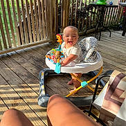 Waylon is registered to the contest to win money with this photo: baby, walker, toy, porch, wooden_floor, sunlight, smiling, outdoor, greenery, person, legs, tattoo, chair, table, plastic_toy, daylight, fence, seat, relaxation, playtime