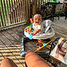 baby, walker, toy, porch, wooden_floor, sunlight, smiling, outdoor, greenery, person, legs, tattoo, chair, table, plastic_toy, daylight, fence, seat, relaxation, playtime